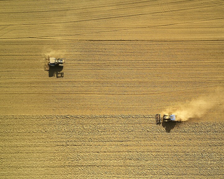 Aerial view of two tractors plowing a large, golden-brown field, creating parallel lines in the soil.